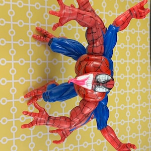 Peut inclure: Figurine articul&eacute;e de Spider-Man, en rouge et bleu, dans une pose dynamique. Le personnage pr&eacute;sente des motifs de toile d'araign&eacute;e sur les parties rouges et des manches bleues. Sur fond jaune.