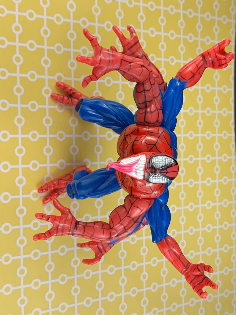 Peut inclure: Figurine articul&eacute;e de Spider-Man, en rouge et bleu, dans une pose dynamique. Le personnage pr&eacute;sente des motifs de toile d'araign&eacute;e sur les parties rouges et des manches bleues. Sur fond jaune.