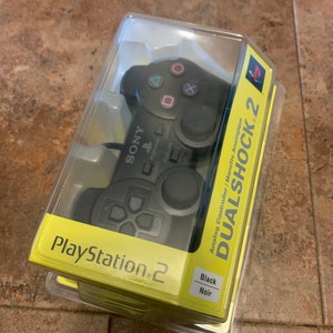 Peut inclure: Une manette noire PlayStation 2 DualShock 2 dans son emballage d'origine. La manette est dotée de joysticks analogiques et de plusieurs boutons. L'emballage est jaune avec le logo PlayStation 2 et les mots "DualShock 2" en gros caractères.