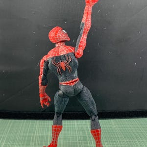 May include: A Spider-Man action figure in a dynamic pose, with one arm raised, holding a small, light-colored object. The figure is predominantly red and black, with a spider emblem on the back. The figure is on a green surface.