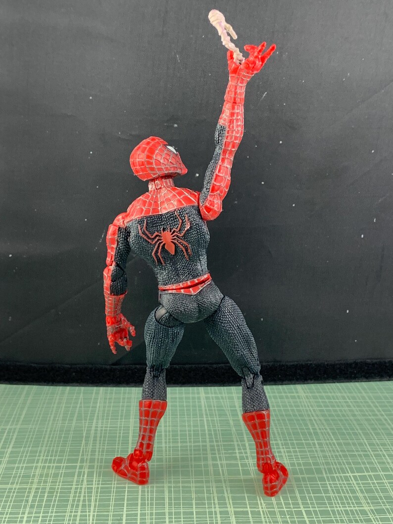 May include: A Spider-Man action figure in a dynamic pose, with one arm raised, holding a small, light-colored object. The figure is predominantly red and black, with a spider emblem on the back. The figure is on a green surface.