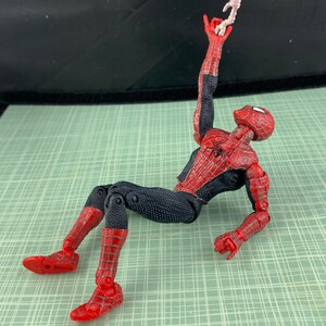May include: A Spider-Man action figure in a dynamic pose, featuring a red and black costume. The figure is holding a small, off-white object in its raised hand. The figure is lying on a green surface.