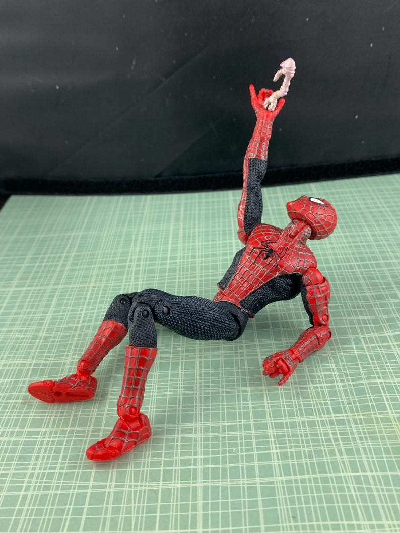 May include: A Spider-Man action figure in a dynamic pose, featuring a red and black costume. The figure is holding a small, off-white object in its raised hand. The figure is lying on a green surface.