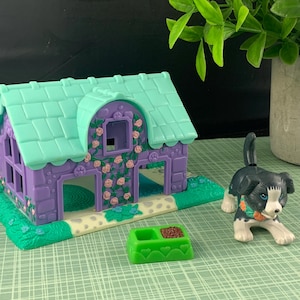 Könnte beinhalten: Ein Miniatur-Puppenhaus in Lila und Grün mit passendem Hund und Futternapf. Das Puppenhaus hat ein grünes Dach und ist mit rosa Rosen verziert. Der Hund ist schwarz und weiß mit einem kleinen orangefarbenen Halsband. Die Futterschale ist grün.
