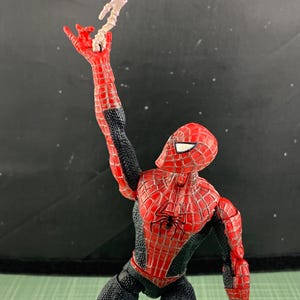 May include: A Spider-Man action figure in a dynamic pose, clad in a red and black suit with web detailing. The figure is holding a white web-like accessory. The figure's pose suggests action and movement, with a detailed spider emblem on the chest.