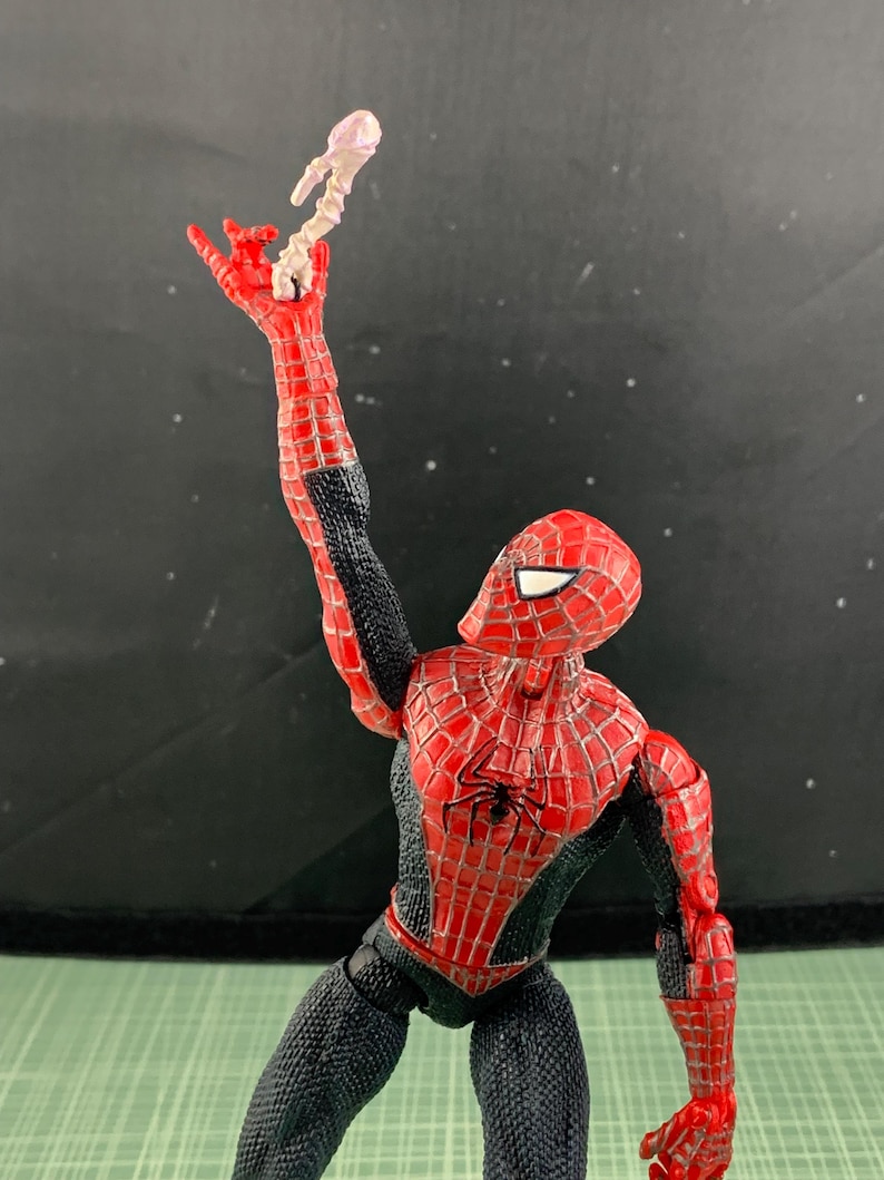 May include: A Spider-Man action figure in a dynamic pose, clad in a red and black suit with web detailing. The figure is holding a white web-like accessory. The figure's pose suggests action and movement, with a detailed spider emblem on the chest.