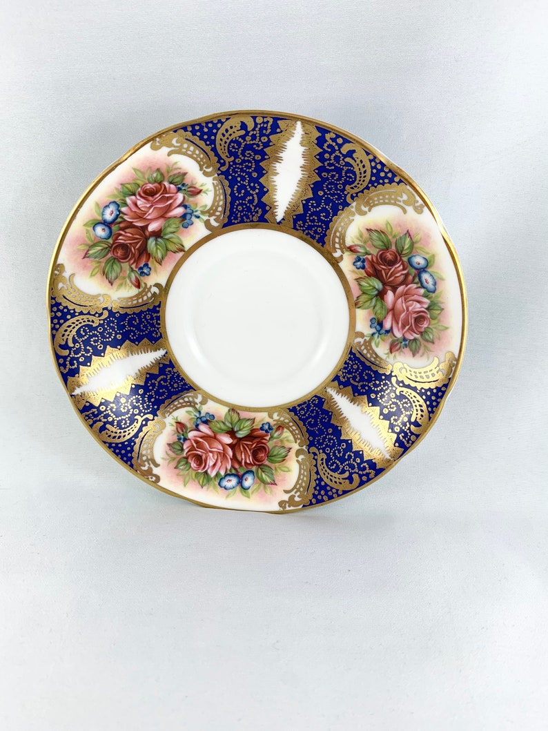 May include: A white porcelain saucer with a blue and gold floral design. The saucer features a delicate floral pattern with pink roses and gold accents.