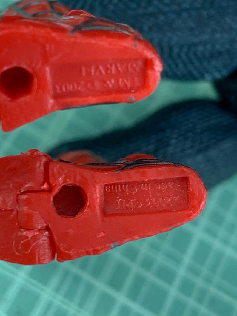 May include: Close-up of two red plastic toy feet. The feet have a hole in the center and the text "TM & &copy; 2003 MARVEL" is visible on one foot. The other foot has the text "Made in China".