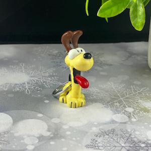 May include: A yellow dog keyring with brown ears and a red tongue. The dog has a black nose and is attached to a silver keyring. The keyring is a cartoon-like design.