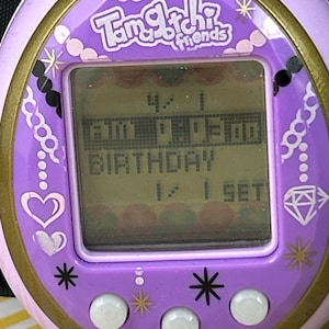 May include: A close-up of a purple Tamagotchi Friends digital pet game. The screen displays the time and the word "BIRTHDAY". The device has a gold trim and decorative elements like hearts, diamonds, and stars.