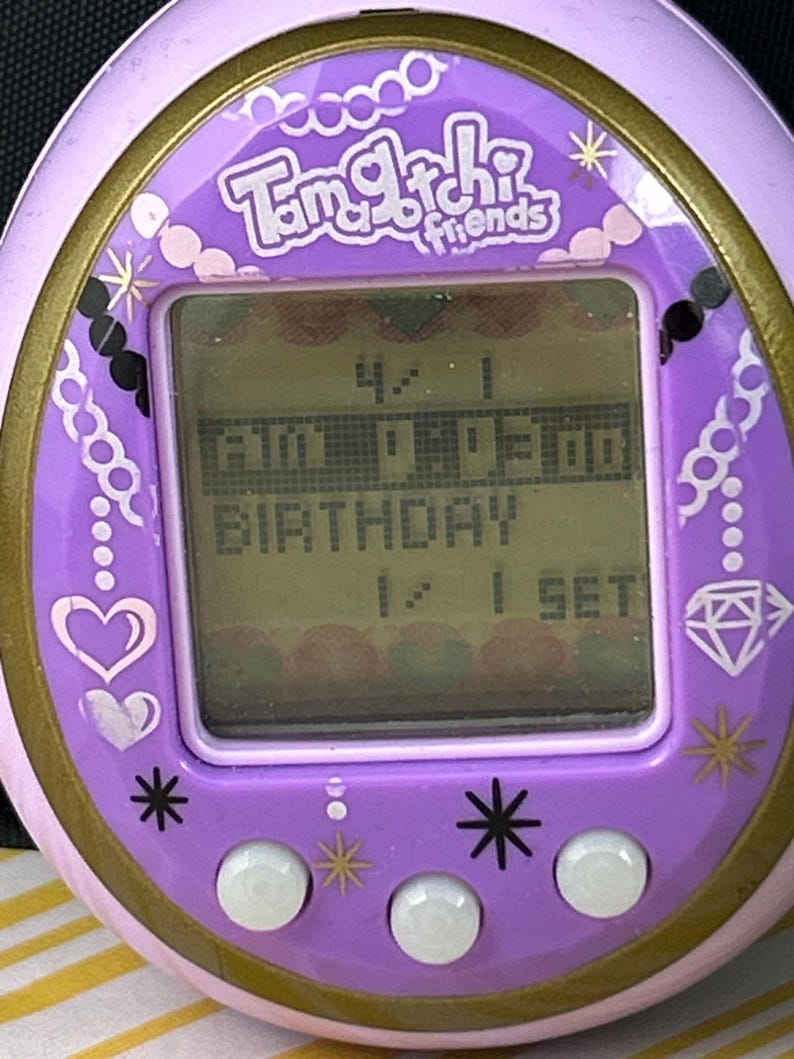 May include: A close-up of a purple Tamagotchi Friends digital pet game. The screen displays the time and the word "BIRTHDAY". The device has a gold trim and decorative elements like hearts, diamonds, and stars.