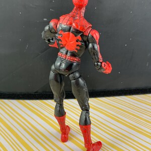 K&ouml;nnte beinhalten: Eine Spider-Man-Actionfigur, von hinten betrachtet, mit rotem und schwarzem Farbschema. Der Kopf und die Stiefel der Figur sind rot, w&auml;hrend der K&ouml;rper schwarz mit roten Netzdetails ist. Ein gro&szlig;es rotes Spinnenemblem befindet sich auf dem R&uuml;cken.