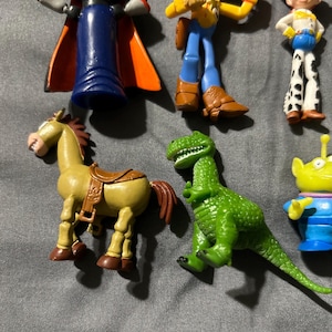 Pu&ograve; includere: Una collezione di figurine colorate dei personaggi di Toy Story, tra cui Buzz Lightyear, Woody, Jessie, un cavallo, un dinosauro e un alieno. Le figure sono di plastica e in varie pose.