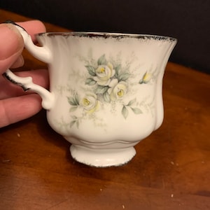 May include: White teacup with a floral design of white roses and green leaves. The cup has a silver rim and a silver handle.
