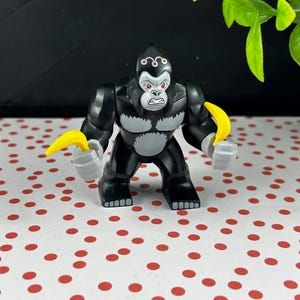 May include: A black and grey gorilla figurine with a white chest and red eyes. The gorilla holds a yellow banana in each hand. The figurine has a detailed design and is standing upright.