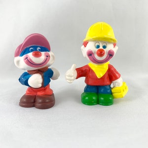 Peut inclure: Deux figurines de clowns colorés. L'un porte un chapeau rose et bleu, une veste bleue et un pantalon marron. L'autre porte un casque jaune, une chemise rouge, un pantalon bleu et des chaussures vertes, et fait un pouce levé.