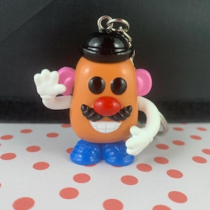 Op de afbeelding: Een Mr. Potato Head sleutelhanger met een lachend gezicht, zwarte hoed, rode neus en een zwarte snor. De sleutelhanger heeft blauwe schoenen, roze oren en witte handen. Het is bevestigd aan een zilveren metalen ring.
