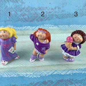 Può includere: Tre figurine Cabbage Patch Kids in abiti diversi. La prima figurina indossa una camicia da notte viola e tiene un libro. La seconda figurina indossa un completo da allenamento viola e bianco con una stella sulla maglietta. La terza figurina indossa un vestito viola e tiene un cono gelato rosa.