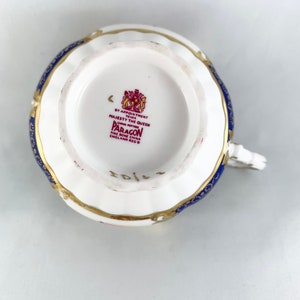 May include: A white teacup with a blue and gold pattern. The bottom of the cup has a red stamp that reads "By Appointment to Her Majesty The Queen China Potters Paragon Fine Bone China England Reg'd".