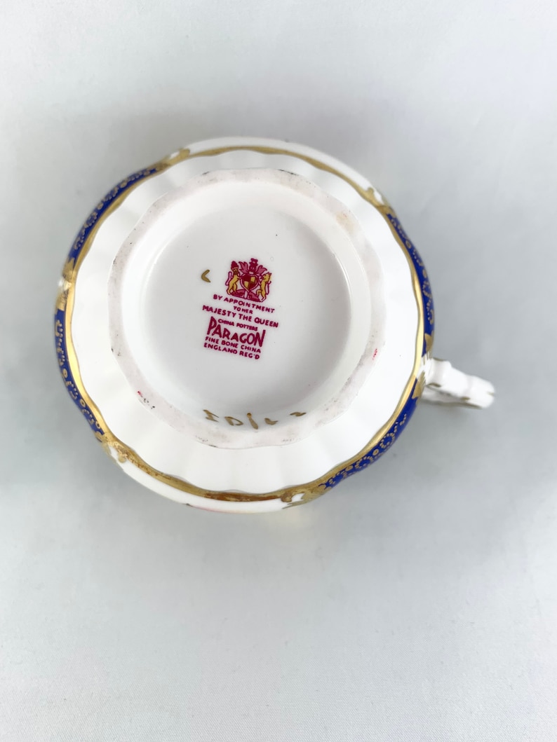 May include: A white teacup with a blue and gold pattern. The bottom of the cup has a red stamp that reads "By Appointment to Her Majesty The Queen China Potters Paragon Fine Bone China England Reg'd".