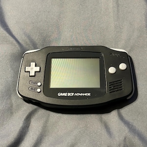 May include: A black Game Boy Advance handheld console with white accents. The console features a screen, directional pad, start and select buttons, and two action buttons. The text "GAME BOY ADVANCE" is printed below the screen.
