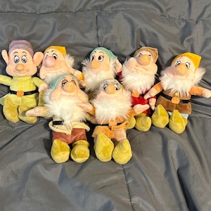 May include: A collection of seven plush dwarf dolls, each with a unique hat and outfit. The dolls have white beards, rosy cheeks, and outstretched arms. The dwarfs are in various colors, including yellow, green, and brown.