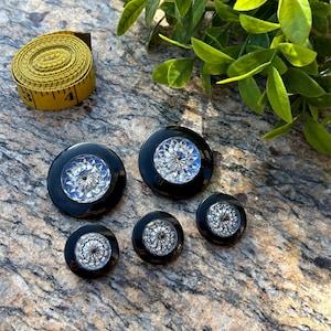 May include: Set of five black and silver buttons with a decorative design. The buttons have a raised, circular design with a clear centre and a black outer ring.