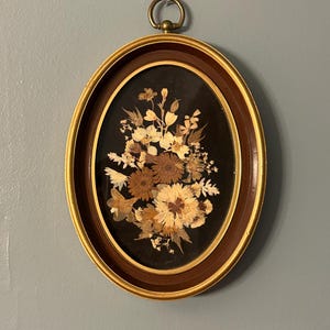 以下が含まれることがあります： 暗い背景の楕円形の額入りフラワーアレンジメント。フレームは、内側に金色の縁と外側に茶色の縁があります。アレンジメントは、白、クリーム、茶色の色合いのドライフラワーが特徴です。真鍮製のリングが上部にあり、吊り下げられます。