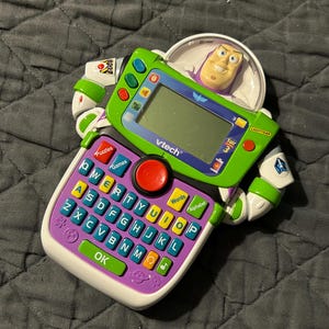 May include: A green and purple VTech Toy Story 3 Buzz Lightyear learning toy. It features a keyboard, screen, and Buzz Lightyear figure. The toy has buttons for games, puzzles, words, and the alphabet.