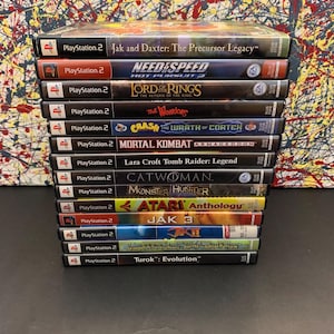May include: A stack of 13 PlayStation 2 video game cases. The games include Jak and Daxter: The Precursor Legacy, Need for Speed: Hot Pursuit 2, The Lord of the Rings: The Return of the King, Crash Bandicoot: The Wrath of Cortex, Mortal Kombat: Armageddon, Lara Croft Tomb Raider: Legend, Catwoman, Monster Hunter, Atari Anthology, Jak 3, Jak II,  and Turok: Evolution.