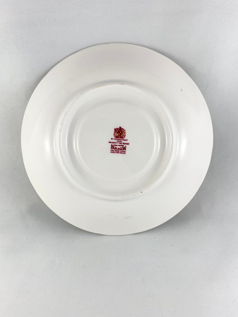 May include: White saucer with a burgundy rim and a burgundy stamp in the centre. The stamp reads "By Appointment to Her Majesty The Queen Paragon Fine Bone China England  Reg'd"