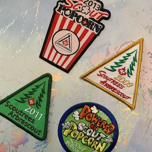 Puede incluir: Cuatro parches bordados que representan un cubo de palomitas de maíz con el texto "Scout Popcorn" y los años 2009, 2011, 2013 y 2009. Los parches son verdes, amarillos y rojos.
