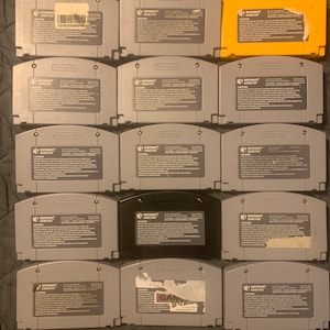 May include: A collection of 12 Nintendo 64 game cartridges, including a yellow cartridge, a black cartridge, and 10 gray cartridges. The cartridges are arranged in four rows of three. The cartridges are all in good condition and have no visible damage.
