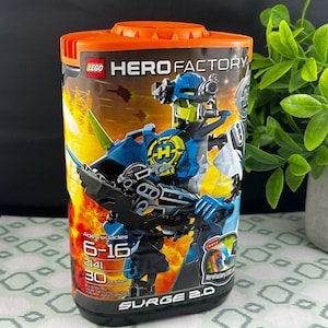 Op de afbeelding: Een cilindrische LEGO Hero Factory set container. De container is oranje en zwart, met een afbeelding van een blauw-zwarte robot. De tekst "SURGE 2.0" is zichtbaar, samen met de leeftijdscategorie "6-16".