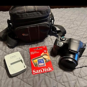 May include: A black Canon digital camera with a lens cap, a black camera bag, a Canon battery charger, and a red SanDisk 32 GB SD card. The camera bag has a shoulder strap.