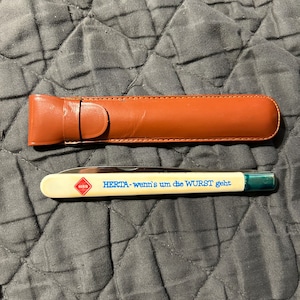 May include: A white pocket knife with a blue handle and a brown leather sheath. The knife has the text "HERTA - wenn's um die WURST geht" printed on it.