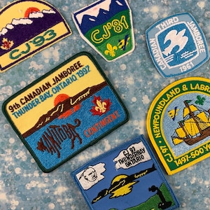 May include: A collection of nine vintage embroidered patches featuring the Canadian Jamboree. The patches are blue, green, yellow, and white with various designs including mountains, a dove, a ship, and a maple leaf. The patches have text that reads "CJ93", "CJ'81", "9th Canadian Jamboree Thunder Bay Ontario 1997", "Third Canadian Jamboree 1961", "CJ 97 Thunderbay Ontario", "Brant District Participant", "CJ 97 Newfoundland & Labrador 1497-500 Y".