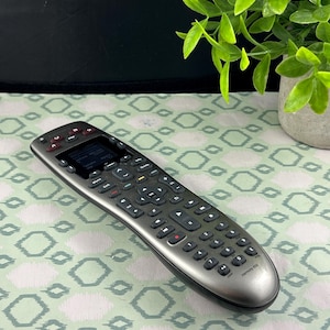 Peut inclure: Une télécommande universelle Logitech Harmony 650 argentée et noire. La télécommande comporte de nombreux boutons et un petit écran. La télécommande est posée sur une surface à motifs avec une plante en arrière-plan.