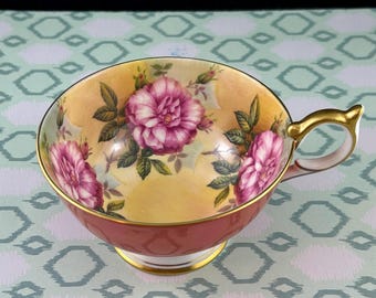 Può includere: Una tazza da tè rossa con un bordo e un manico dorati. L'interno della tazza è decorato con un motivo floreale di rose rosa e foglie verdi su sfondo giallo.