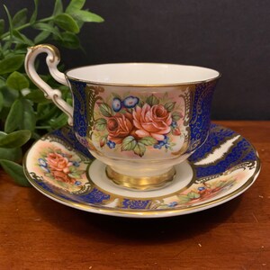 May include: A blue and gold teacup and saucer set with a floral design. The cup and saucer are decorated with pink roses, green leaves, and gold accents.
