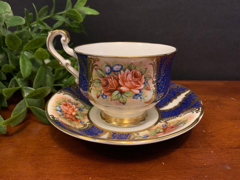 May include: A blue and gold teacup and saucer set with a floral design. The cup and saucer are decorated with pink roses, green leaves, and gold accents.