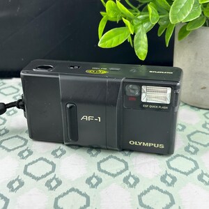May include: Black Olympus AF-1 35mm film camera. The camera has a built-in flash and the text "ESP QUICK FLASH" is visible. The camera also has the text "OLYMPUS" in gold lettering. The camera is on a patterned surface.