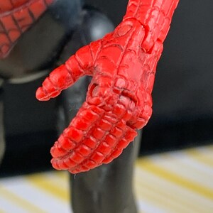 K&ouml;nnte beinhalten: Nahaufnahme einer roten, beweglichen Hand einer Spider-Man-Actionfigur. Die Hand befindet sich in einer entspannten Position, mit sichtbaren Details der strukturierten Oberfl&auml;che und des spinnennetzartigen Musters. Der schwarze Anzug der Figur ist im Hintergrund zu sehen.