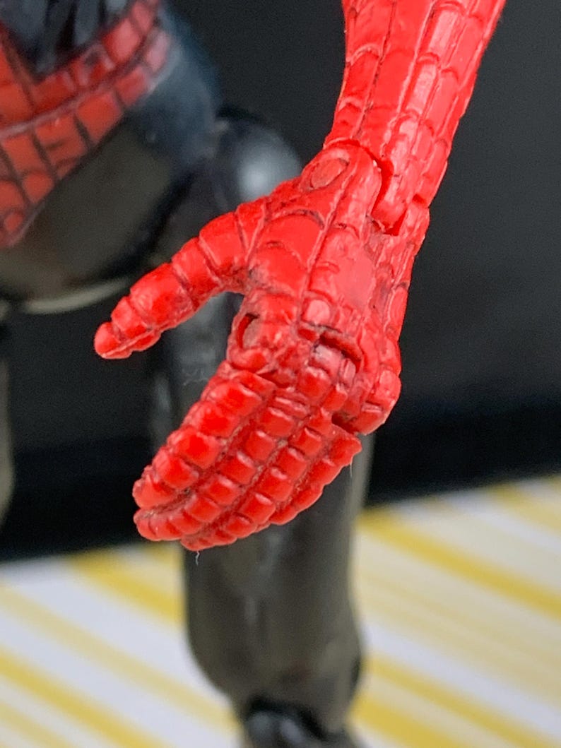 K&ouml;nnte beinhalten: Nahaufnahme einer roten, beweglichen Hand einer Spider-Man-Actionfigur. Die Hand befindet sich in einer entspannten Position, mit sichtbaren Details der strukturierten Oberfl&auml;che und des spinnennetzartigen Musters. Der schwarze Anzug der Figur ist im Hintergrund zu sehen.