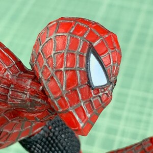 May include: Close-up of a Spider-Man action figure's head and shoulder. The figure features a red and black costume with a webbed pattern on the head. The eyes are white, and the figure is posed in a dynamic position.