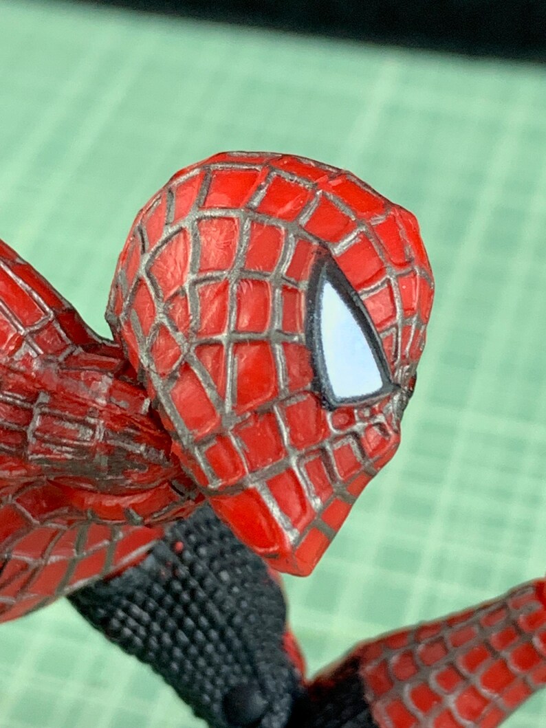May include: Close-up of a Spider-Man action figure's head and shoulder. The figure features a red and black costume with a webbed pattern on the head. The eyes are white, and the figure is posed in a dynamic position.