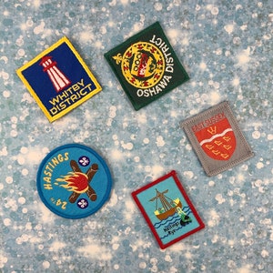 May include: Four colourful embroidered patches with different designs. The patches are square and round, with yellow, blue, red, green and grey colours. The patches have text that reads "Whitby District", "Oshawa District", "Hastings 24", "East Sussex" and "Hastings Rye".