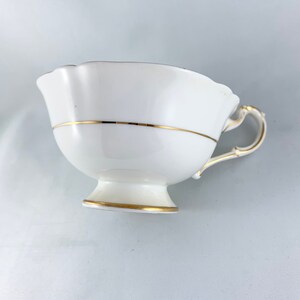 K&ouml;nnte beinhalten: Wei&szlig;e Teetasse mit goldener Verzierung am Rand und am Boden. Die Teetasse hat einen gewellten Rand und einen zarten Henkel.