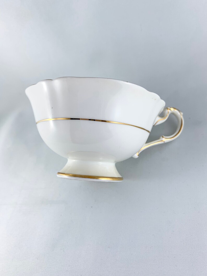 K&ouml;nnte beinhalten: Wei&szlig;e Teetasse mit goldener Verzierung am Rand und am Boden. Die Teetasse hat einen gewellten Rand und einen zarten Henkel.