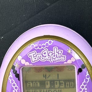May include: A purple Tamagotchi Friends digital pet toy with a gold trim. The screen displays the time and the word "BIRTHDAY". The device has decorative elements including hearts and jewels.